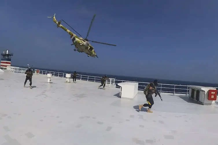 Armed men on the deck of a ship while a helicopter flies above.