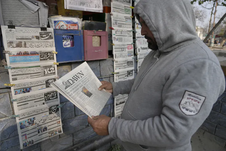 A man in a grey hoodie holds a newspaper as he reads the front page.