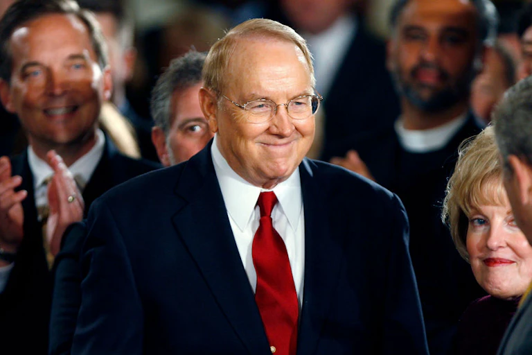 A man with sandy-colored hair, glasses, a red tie and a blue suit walks smiling through a crowd of people.