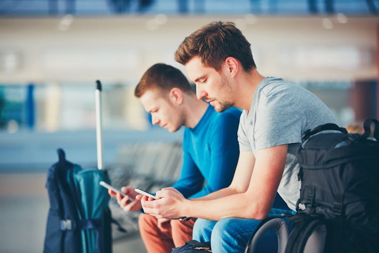 two men sit on a bench, both looking at their mobile phones
