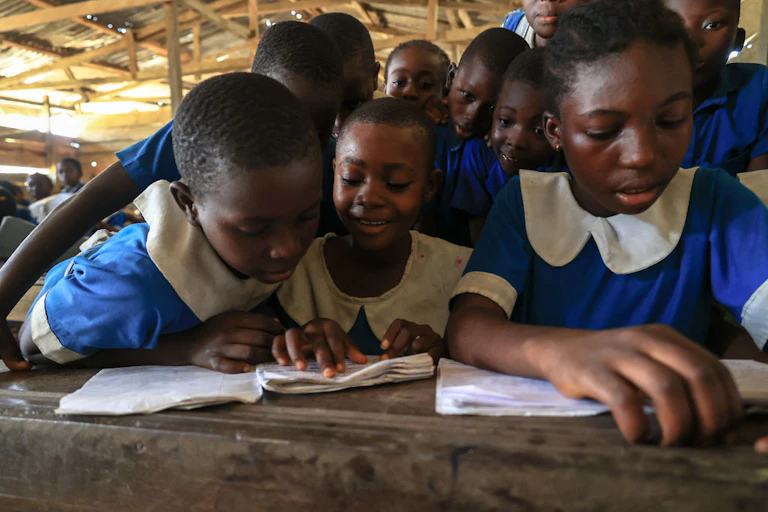 A group of young girls in blue and white school uniforms crowd around a desk to read together from two books