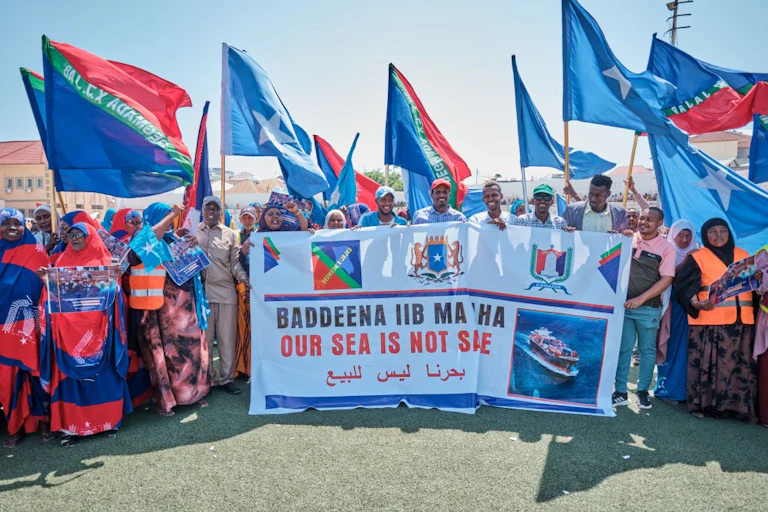Men and women holding up blue flags with a star in the middle, and striped blue, green and red flags while holding a banner written 'Our Sea is not Safe'