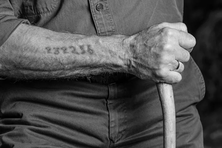 A black and white photograph of an old man with a number tattooed on his forearm.