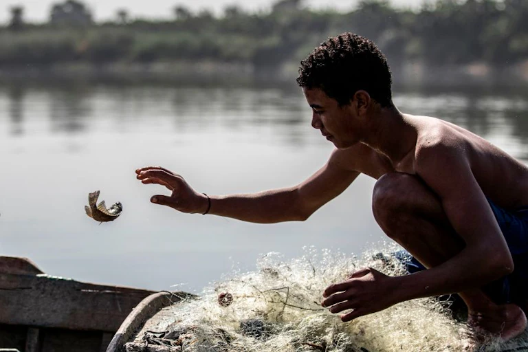 A young man, who is shirtless and wearing shorts, reaches for a fish that's flopping, seemingly halfway in the air, above his net.