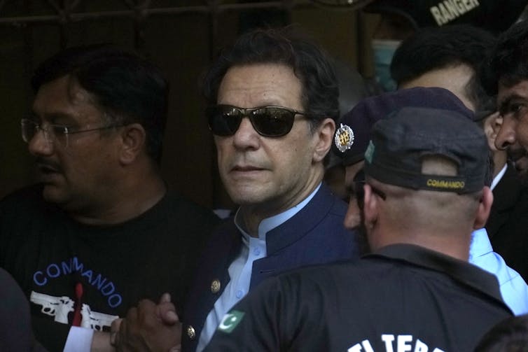 A man in sunglasses smiles faintly surrounded by security guards.