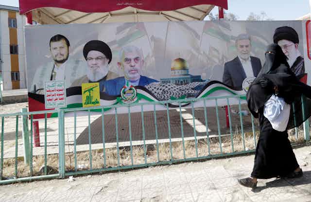A woman in an Islamic dress walks past a billboard with various men on it.
