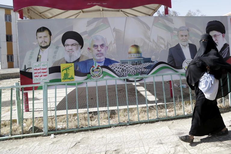 A woman in an Islamic dress walks past a billboard with various men on it.