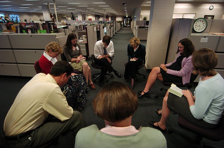 Eight adults in business attire sit on chairs in a circle, heads bowed, in the middle of an open-floor office.
