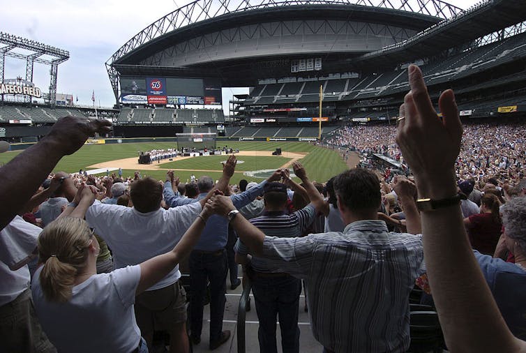 Rows of people hold hands and raise them up as they assemble in a baseball stadium.