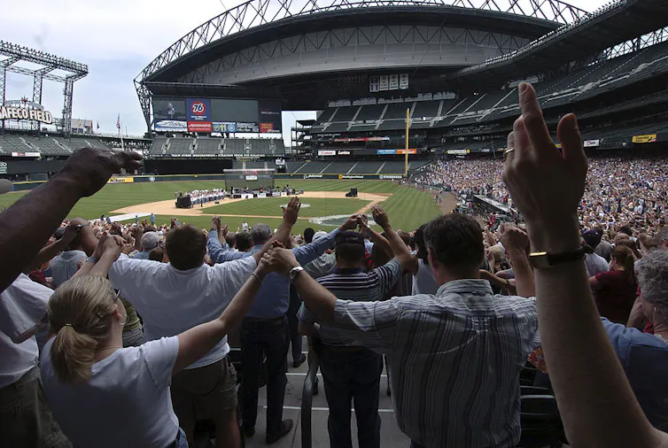 Rows of people hold hands and raise them up as they assemble in a baseball stadium.