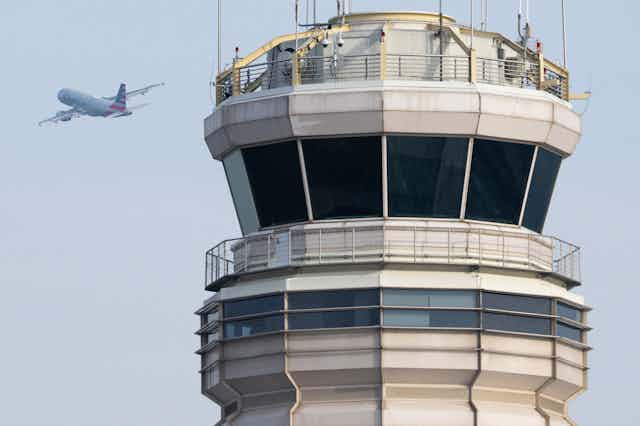 A glass-walled room at the top of a tower with a jet liner in the air in the background