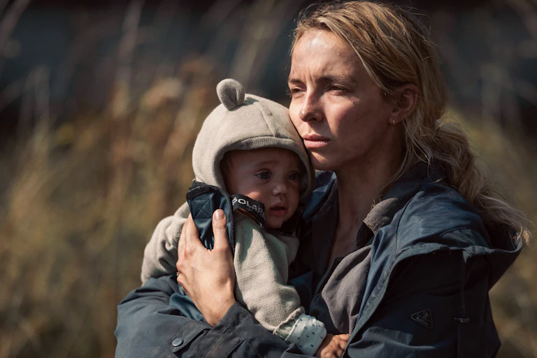 Woman looks distressed as she holds baby close, outside in grassy area.