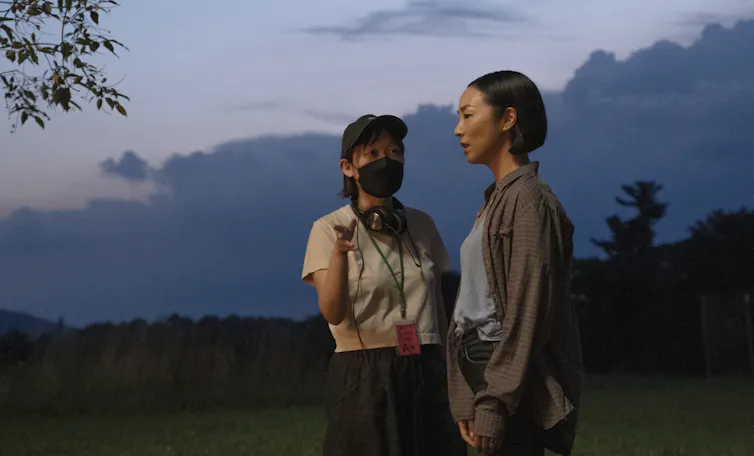 A woman in a face mask seen talking to a taller woman staring in the distance.