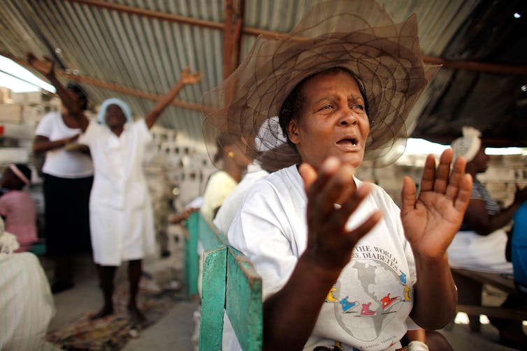 A woman in a wide-brimmed hat holds her hands up as she prays, with some other people in the background.