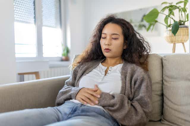A woman with cramps sits on a couch while holding her stomach in pain.