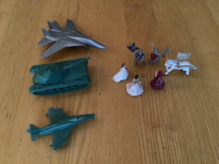 On a tabletop, small figurines of princesses, knights and horses are gathered in a circle, while a miniature tank and two miniature planes are pointed at them.