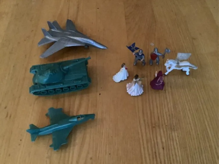 On a tabletop, small figurines of princesses, knights and horses are gathered in a circle, while a miniature tank and two miniature planes are pointed at them.