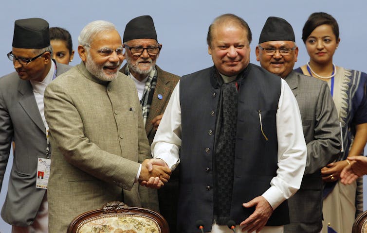 Two men, one in a collarless jacket, shake hands and smile.