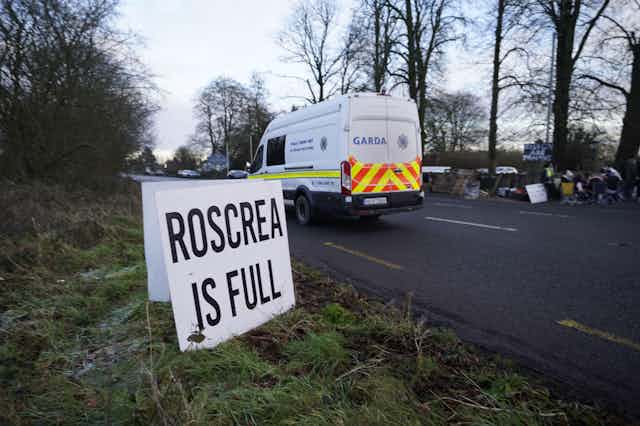 A sign on the side of a road that reads Roscrea is full. A Garda van is driving past.