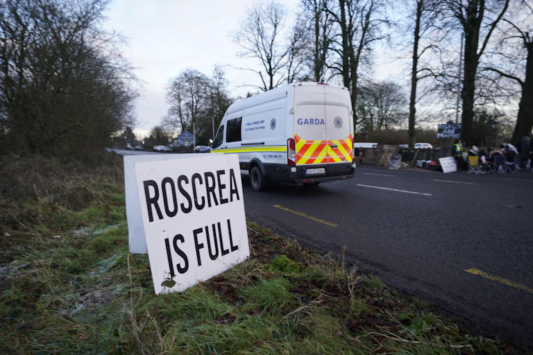 A sign on the side of a road that reads Roscrea is full. A Garda van is driving past.