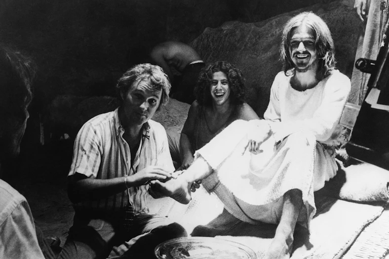 A man in a striped open shirt holds the foot of a man in a Jesus-like white robe and beard.