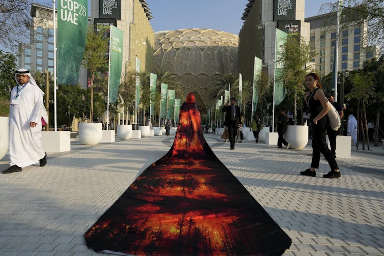 A woman wearing a dress with a long train with images of forest fires walks past other people and buildings.