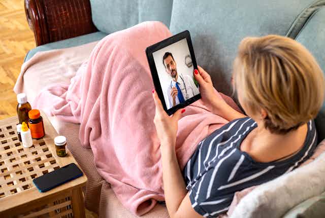 A woman sits on a couch with a blanket over her lap, holding an ipad and talking to her doctor during a telehealth session.