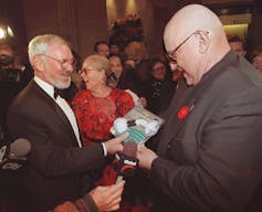 A man with a grey beard in a tux smiles as another man hands him a gift.