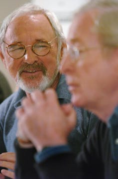 A man with a grey beard and glasses smiles as he listens to another man speak.