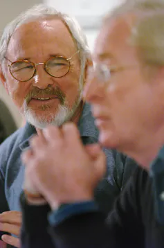 A man with a grey beard and glasses smiles as he listens to another man speak.