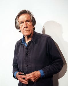 Expressionless elderly man with long gray hair holding a pair of eyeglasses.