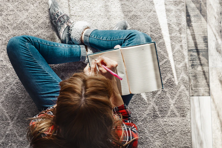 View from above of a young woman sitting cross legged on the floor, writing in a journal