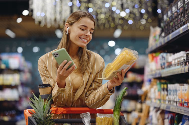 A young woman pushes a grocery cart, smiling and holding her phone in one hand and a bag of pasta in the other