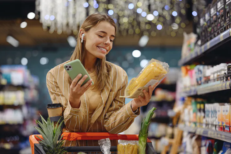 A young woman pushes a grocery cart, smiling and holding her phone in one hand and a bag of pasta in the other