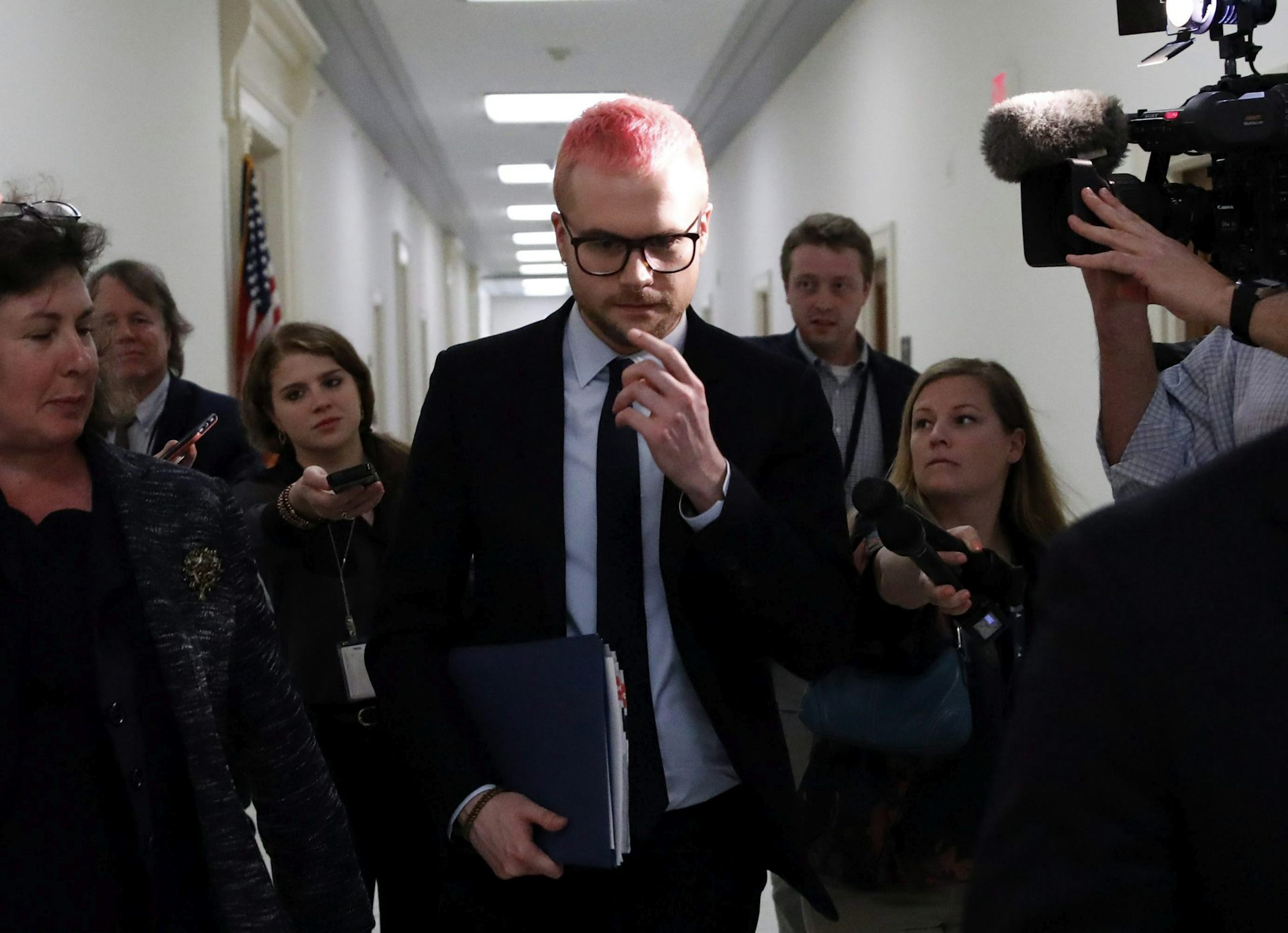 A man with a dyed pink buzzcut and glasses, wearing a suit, walks through a crowd of people down a hallway