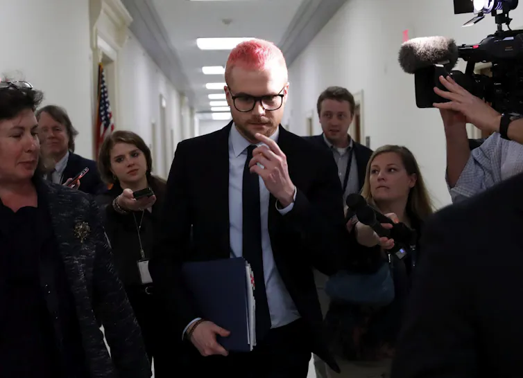 A man with a dyed pink buzzcut and glasses, wearing a suit, walks through a crowd of people down a hallway