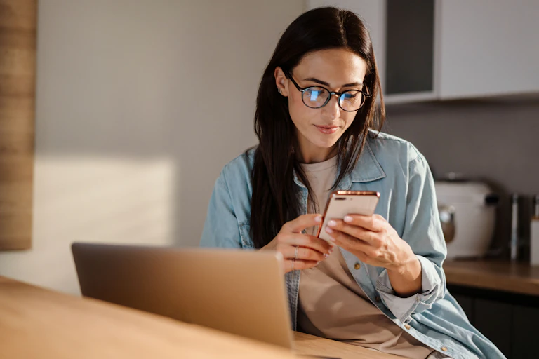 A woman looking at her phone while sitting in front of a laptop at a kitchen table