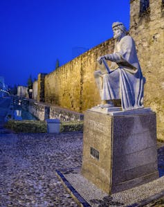 A statue of a seated man in robes on a pedestal, in front of a brightly lit stone wall at night.