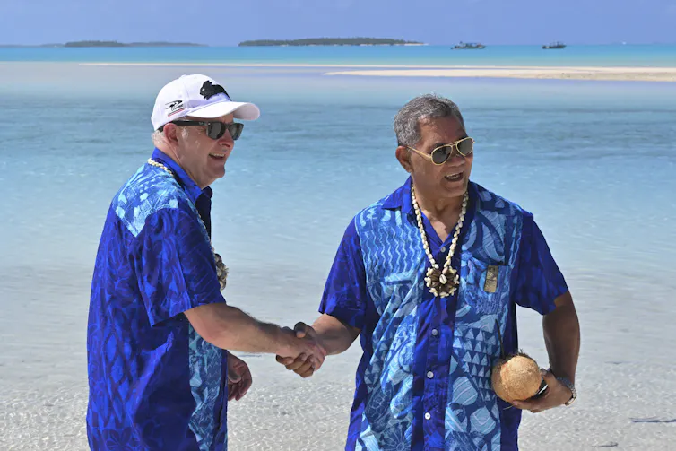 Two men in blue shirts stand on a beach shaking hands