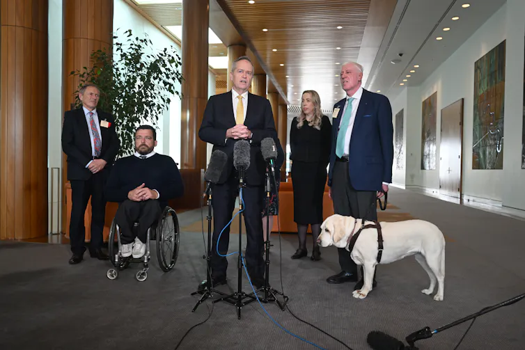 group of people in formal building include one man in wheelchair and a man standing with his guide dog.