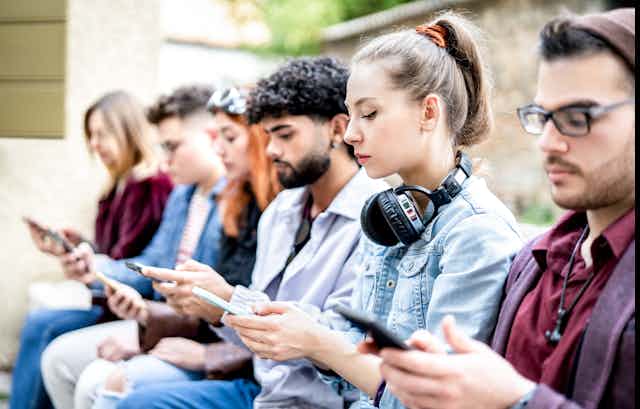 A group of young people sit on a bench as they view and text on their smartphones.