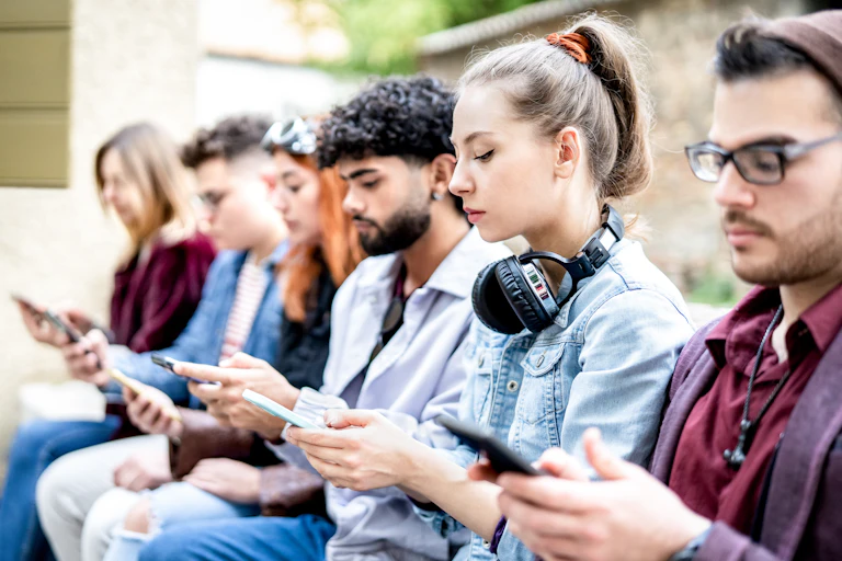 A group of young people sit on a bench as they view and text on their smartphones.