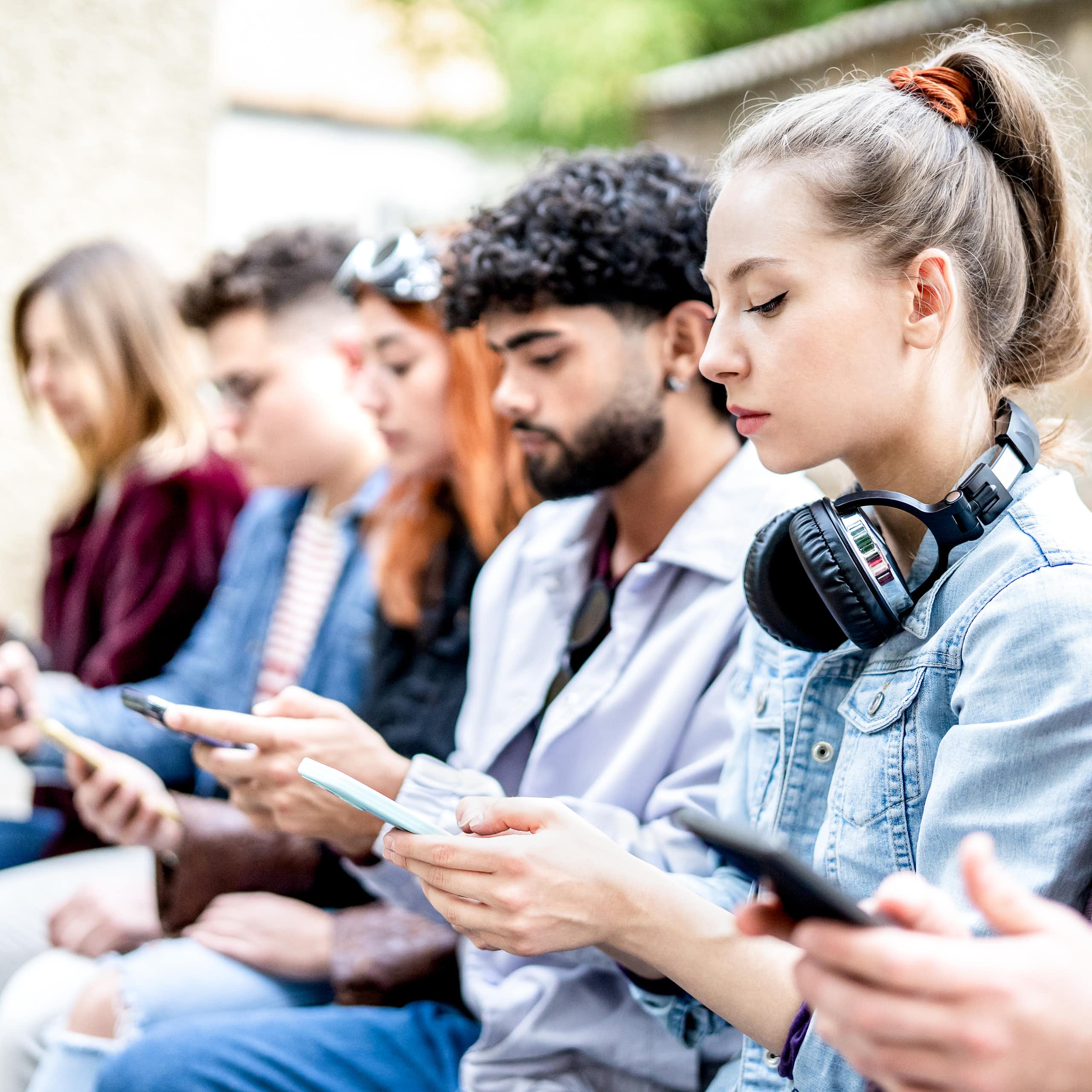 A group of young people sit on a bench as they view and text on their smartphones.