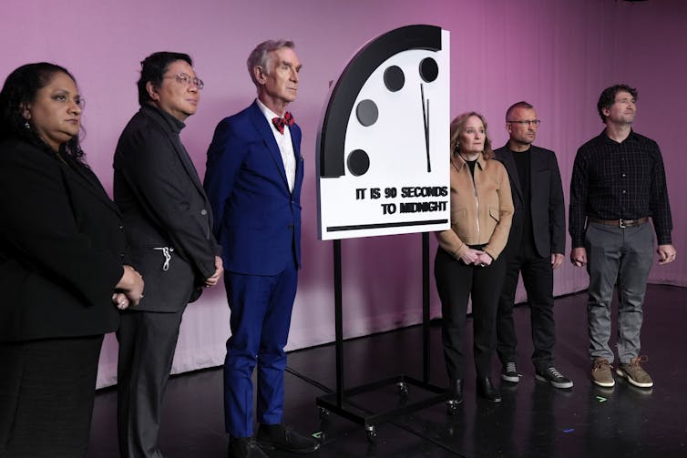 A group of people standing next to a quarter slice of a clock that states it is 90 seconds to midnight