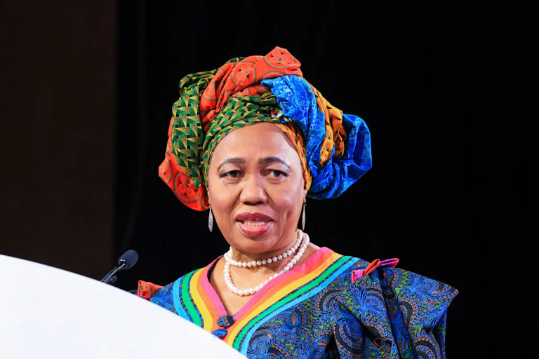 A woman wearing a bright blue, red, green and yellow headdress with a matching dress speaks into a microphone behind a podium