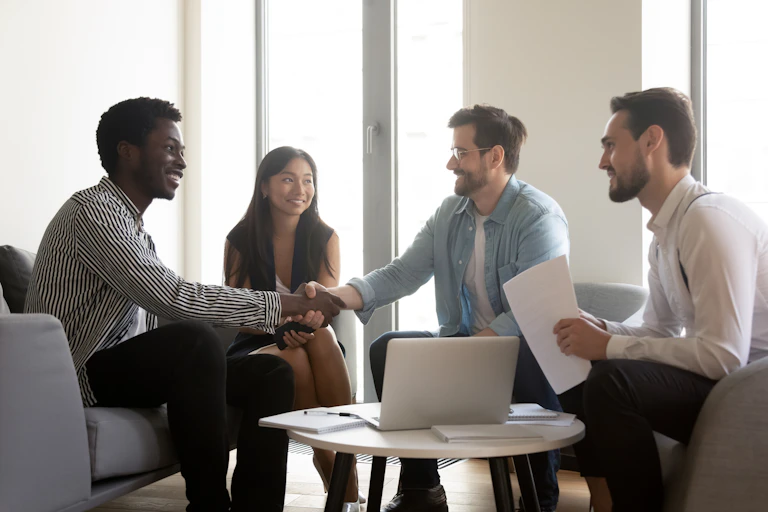 Four employees of different races, smiling and shaking hands around a table