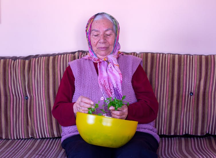 An elderly woman inspects greens in a plastic bowl.