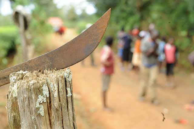 A machete is stuck on a wood stump with people in background
