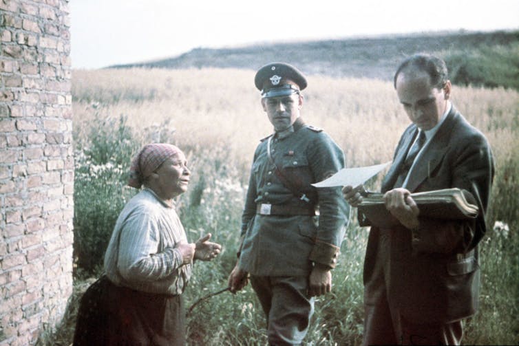 A faded photograph of a soldier and a man in a suit standing as they interview a shorter woman in a kerchief.