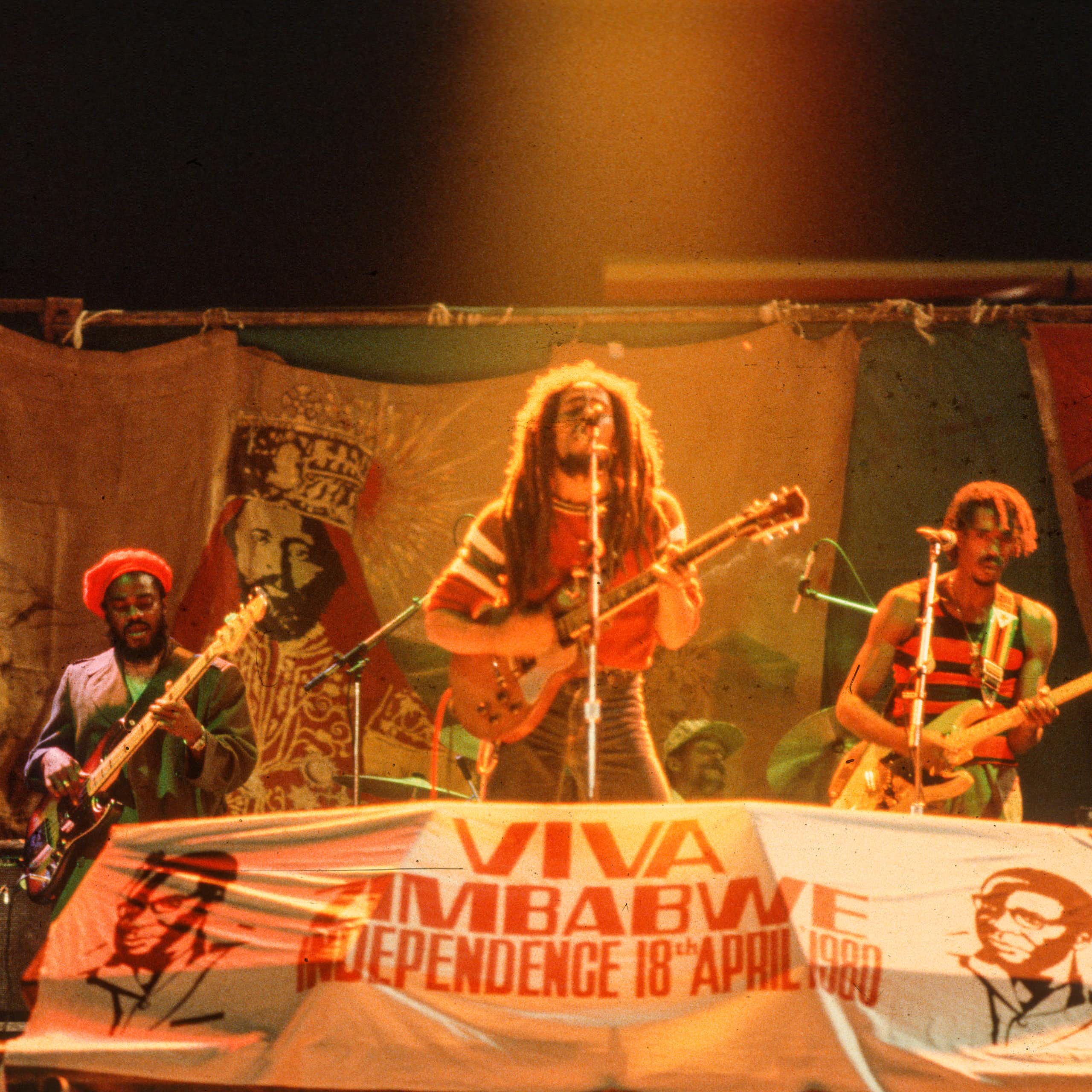 Dreadlocked man sings and plays guitar in front of sign reading 'Viva Zimbabwe.'